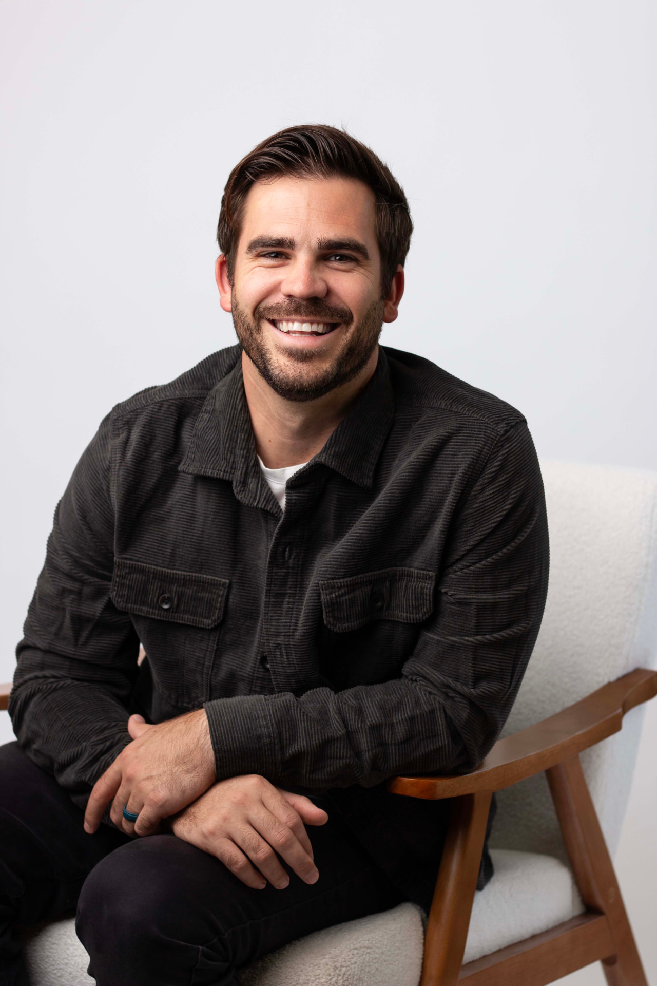 Cortland Watson seated in a wood and cream armchair, wearing a dark gray corduroy shirt jacket over a white t-shirt