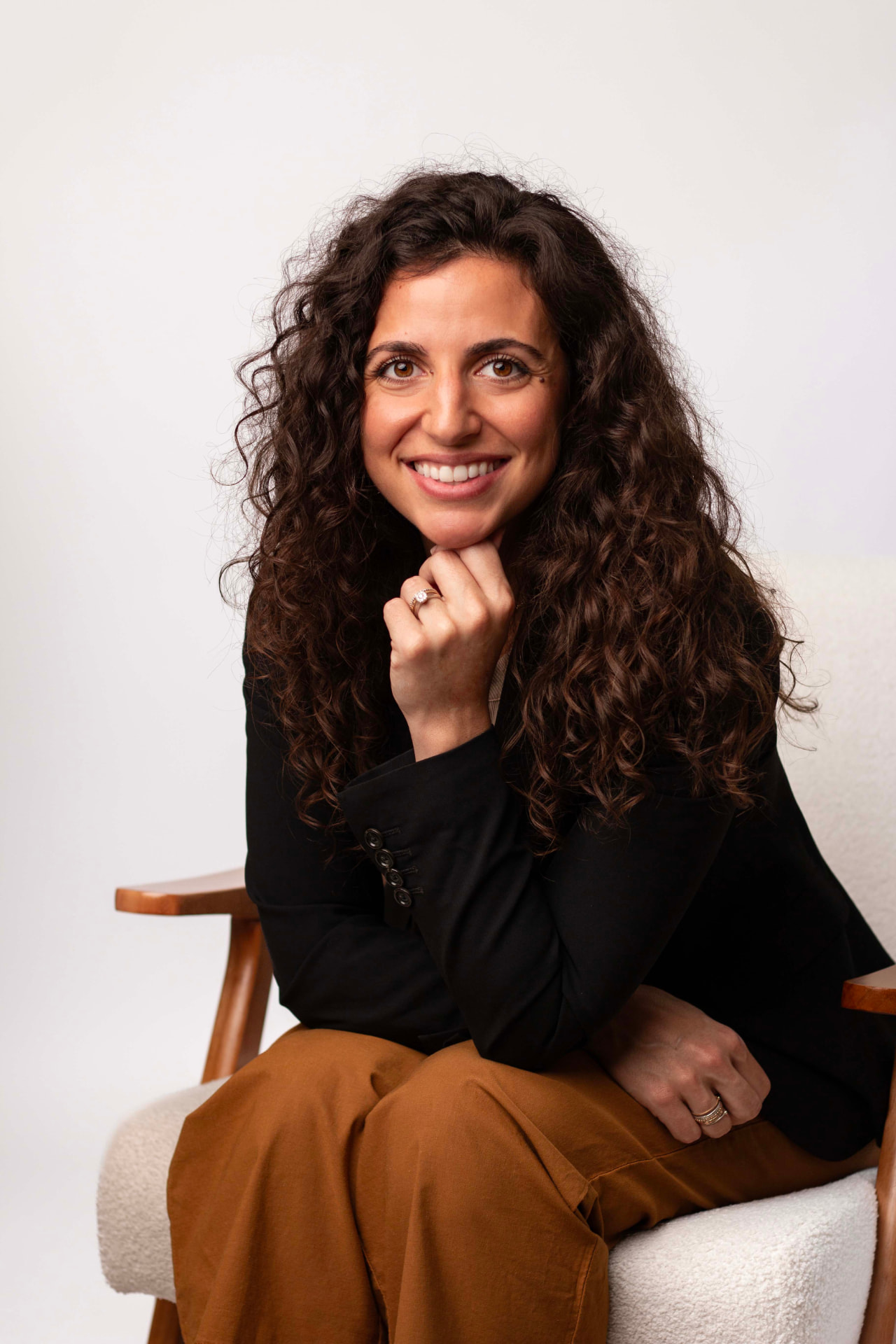 Simone Donae seated in a wood and cream armchair, wearing a black blazer over an off-white top and caramel trousers, against a light gray studio backdrop